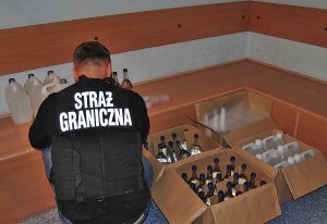 Fot. MOSG Funkcjonariusz Straży Granicznej przy szklanych butelkach i plastikowych pojemnikach z cieczą.